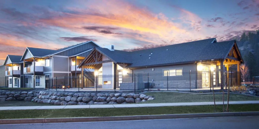 A modern building with wooden beams and large windows is illuminated against a vibrant sunset sky.