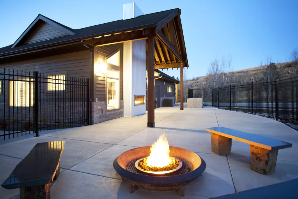 Fire pit with a burning flame on a patio outside a modern house in the early evening, with benches and fencing nearby.
