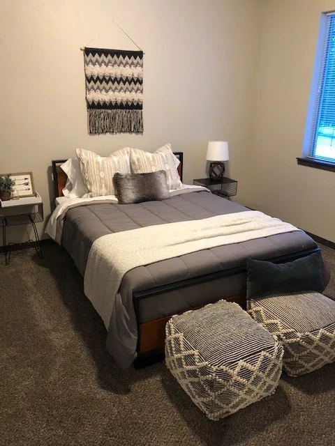 A neatly made bed with gray and white bedding, two patterned ottomans, and a geometric wall hanging in a cozy bedroom.