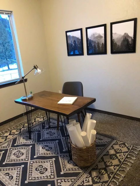 A minimalist home office with a wooden desk, a chair, a lamp, and a basket holding rolled papers on a patterned rug.